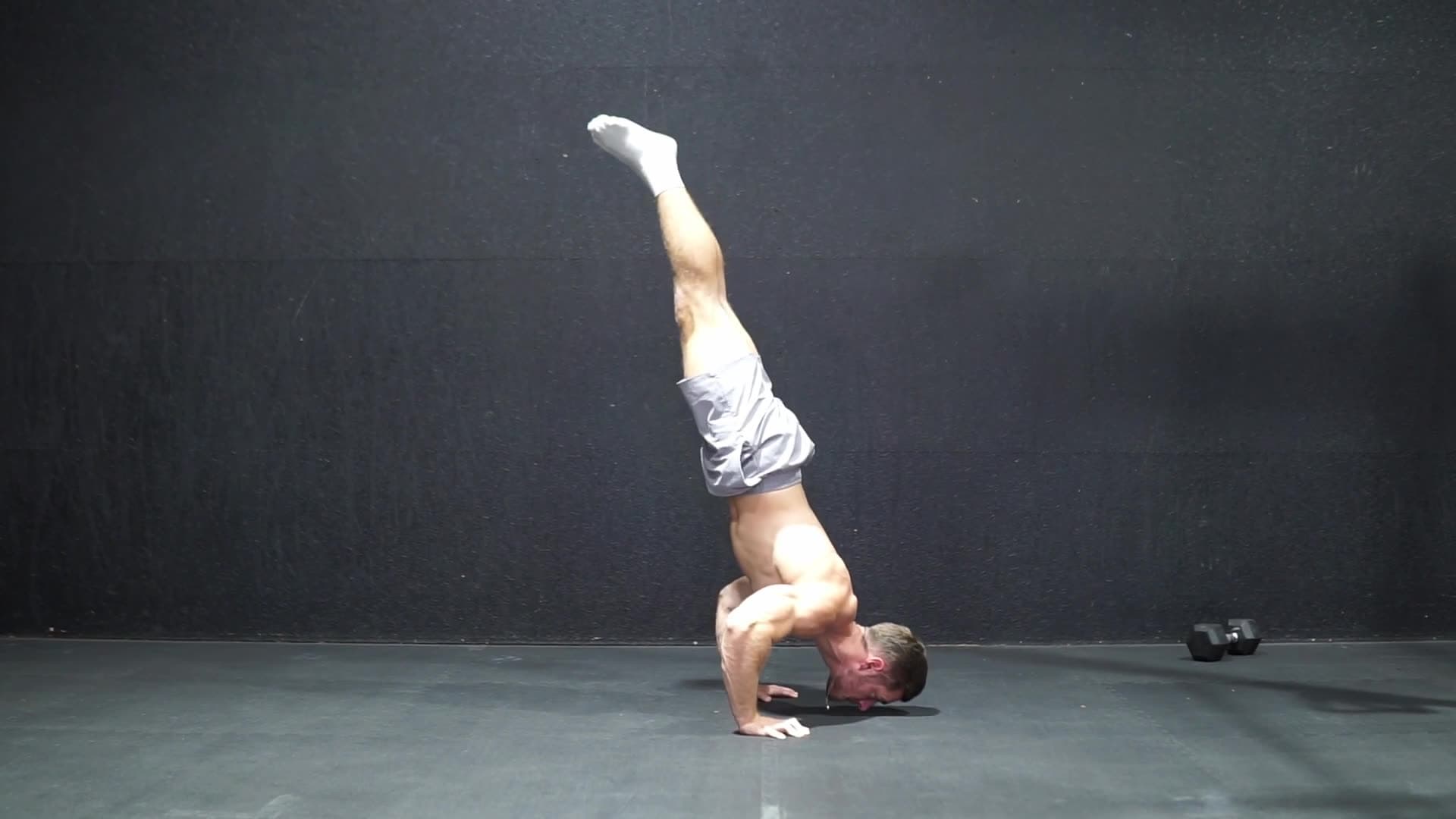 Male athlete at the bottom of a wall-supported handstand push up, head close to the floor with arms bent under full body weight, feet against a concrete wall